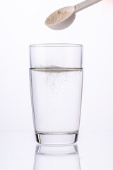 Collagen powder is poured with a measuring spoon into a glass of water on a white background with a reflection. The medicine is diluted in water. Vertical photo