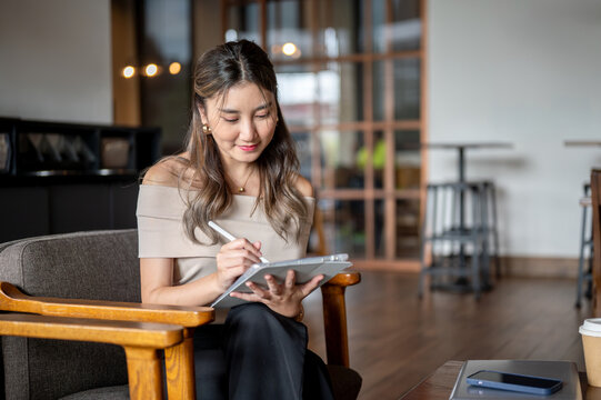 Asian woman writing or drawing in her tablet with a stylus on a wooden chair in cafe or coffee shop.