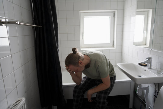 Depressed man sitting at edge of bathtub in bathroom