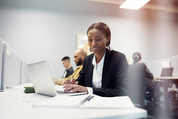 Young female business professional working on laptop while reading details in diary at office