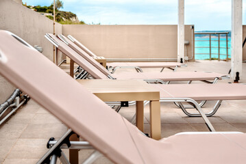 Fototapeta premium Row of beige sun loungers and tables along tiled balcony facing the sea. Modern outdoor seating designed for group relaxation under open sky. Clean design and tranquil beach setting.