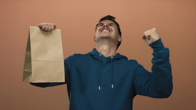 Young hispanic man celebrates holding paper bag against isolated brown wall background, expressing excitement.