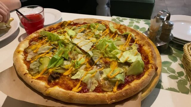 Large pizza Burger with lettuce, beef, cucumbers and cheese spins on table in cafe. Drink is visible in background, along with a basket and condiments.