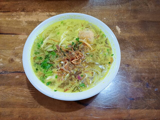 A bowl of Soto Ayam, a traditional Indonesian yellow chicken soup, served on a wooden table. The soup has a creamy, yellow-greenish broth, characteristic of the turmeric and coconut milk