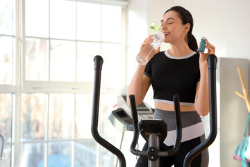 Fototapeta premium Sporty young woman with inhaler and water bottle on exercising bike in gym