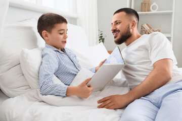 Happy father with his son in pajamas reading book on bed at home