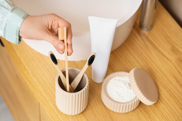 Young woman with toothpaste and powder taking brush from holder near sink in bathroom, top view