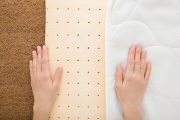 Young adult woman hands touching brown coconut coir, yellow rubber foam and white cloth cover of orthopedic mattress. Showing inside layers. Closeup. Top down view.