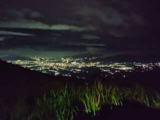 City lights sparkle beneath a dark sky, viewed from a quiet hilltop under clouds at midnight.