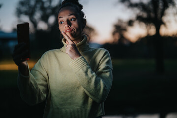 A young girl wearing a yellow sweater captures a playful selfie outdoors during dusk. The dim...