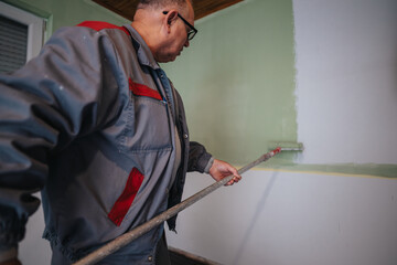 An older man applies light green paint on a wall with a roller, showing a home renovation or...
