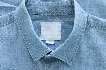 A close-up of a light blue, polka-dotted shirt, focusing on the collar with a blank white label, a button on the placket, and a portion of the sleeve. .