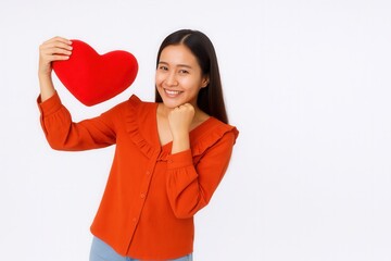 A woman smiles and holds a red heart-shaped cushion, expressing joy and affection, dressed in an orange blouse