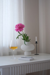 A chic vase of vibrant pink peony, a refreshing wine drink, copybook with pen, and an elegant candle holder in a serene indoor setting