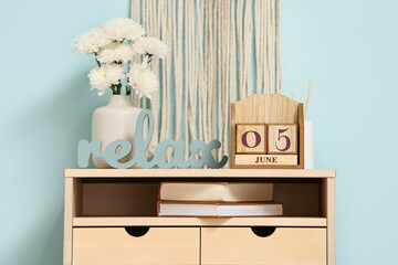 Wooden cube calendar with date 5 June, vase of flowers and books on commode near blue wall in...