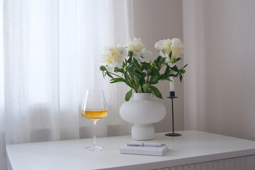 A chic vase of vibrant white peonies, a refreshing wine drink, copybook with pen, and an elegant candle holder in a serene indoor setting