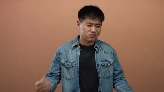 Young man expressing skepticism with hand gestures against an isolated brown wall background, conveying uncertainty and contemplation while wearing a casual denim jacket.