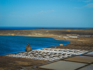 Salt pans of Salinas de Janubio along a stunning blue lagoon on volcanic coast. White mounds of salt line the fields against the Atlantic Ocean backdrop. Traditional sea salt harvesting at Lanzarote.