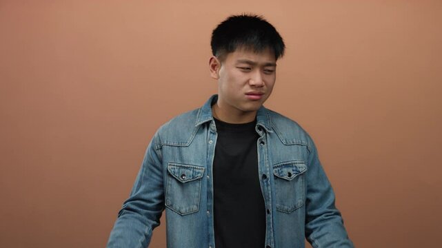 Young man expressing skepticism with hand gestures against an isolated brown wall background, conveying uncertainty and contemplation while wearing a casual denim jacket.