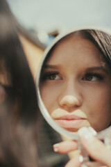 A young woman checks her appearance using a mirror, seemingly outdoors. The light emphasizes her features and the scene gives off a modern and fashionable vibe