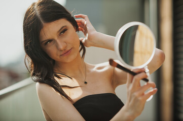 On a balcony in the late afternoon, a young woman wearing a black strapless top holds a mirror and applies makeup. She appears to be getting ready to go out or making a Social Media post