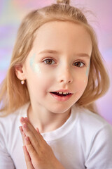 A young girl with long blonde hair and fair skin opened her mouth in surprise. She has glitter on her cheeks, which gives a shiny effect. The girl folded her hands in front of her.