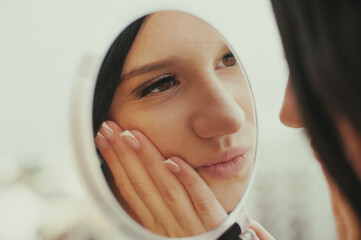 A young woman with black hair gazes into a hand mirror, touching her face. She is possibly checking her appearance or posing for social media, displaying a fashionable look