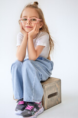 A cute young girl with long blonde hair and pink glasses sits on a wooden box and looks at the camera with a smile, resting her chin on her hands. She is wearing black boots with bright pink laces.