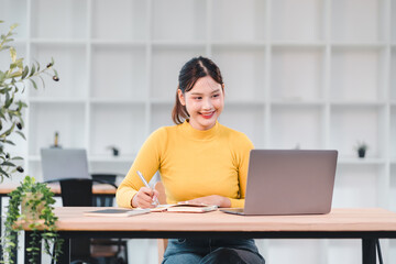 Young Asian woman in yellow sweater working at desk with laptop, smiling and taking notes in modern office with white shelves, plants, and natural light, showing productivity and focus