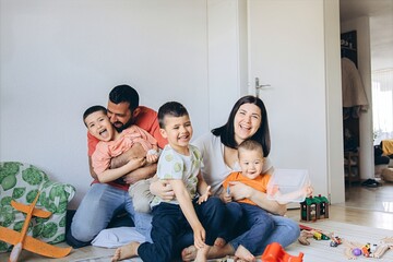 A family with three young children sits together on the floor of bright playroom.Toys,building blocks,and toy airplane are scattered around,creating cozy,playful atmosphere of everyday family life.