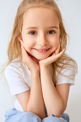 A charming young girl with long blond hair smiles radiantly, resting her head on her hands. She is dressed in a white T-shirt and light trousers. There are delicate blue patterns on her cheeks