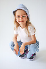 A mischievous girl in a denim panama hat and glasses sits on her haunches and smiles. She is wearing a white T-shirt, blue pants and dark sneakers with pink elements. The child radiates positivity