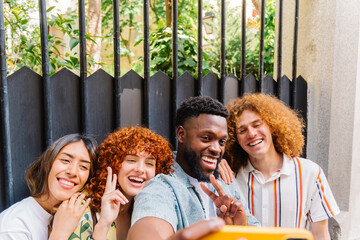 Multiracial friends smiling and flashing peace signs while capturing a selfie outdoors in front of...