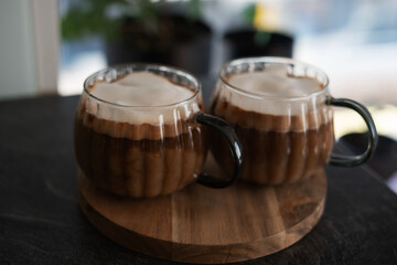 two glass mugs filled with frothy creamy coffee on wooden coaster in softly lit home setting for cozy morning ritual, aesthetic, presentation, espresso, latte, brown, texture, enjoyment, relaxing