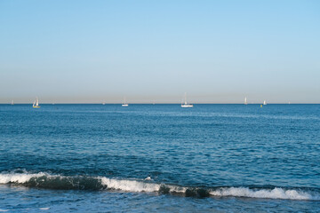 Tranquil seascape featuring calm blue waters, gentle waves lapping at the shore, and sailboats gracefully floating in the distance under a clear sky, evoking a serene coastal atmosphere