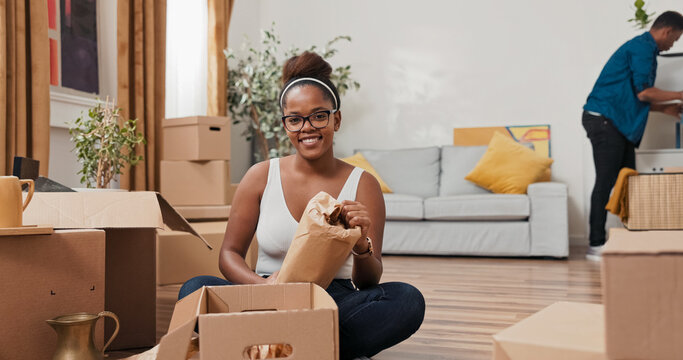 A girl with glasses sits in the middle of her new flat, unpacking belongings. She holds a vase and smiles into the lens, while the man in the background continues cleaning the space.