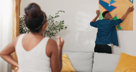 A couple in love decorates their first apartment. The man hangs a painting on the wall while the...