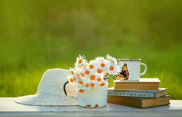 summer nature background. chamomile flowers in mug, butterfly, books and braided hat close up on table in garden. Beautiful rustic floral composition. relaxation, harmony atmosphere. soft focus