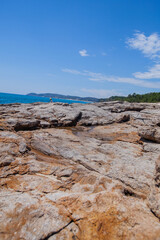  A wide-angle view of a rugged, textured rocky coastline stretching into the foreground, leading to the calm turquoise sea and a clear blue sky in the distance.