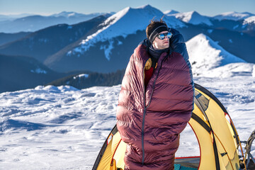Man standing outside tent in snowy mountains, wrapped in sleeping bag, warming up in cold morning...