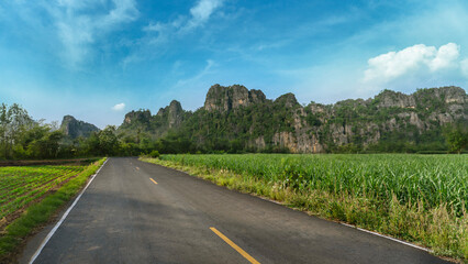 An empty rural road leads through green fields towards limestone mountains under a bright blue sky. Peaceful nature scene perfect for travel, freedom, and adventure themes.