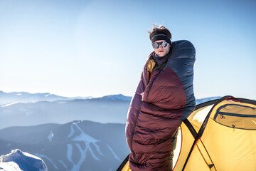 Man standing outside tent in snowy mountains, wrapped in sleeping bag, warming up in cold morning...