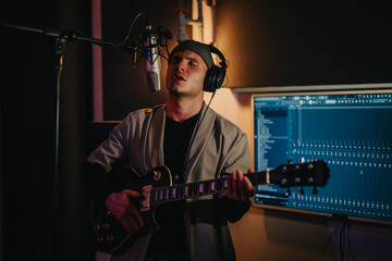 A focused musician performs with a guitar and microphone in a recording studio. He wears headphones...