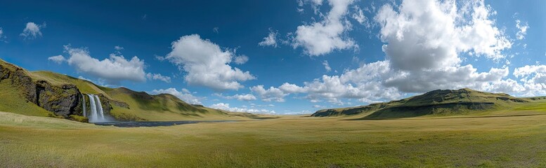 Panoramic view of a waterfall cascading into a valley, surrounded by hills and a vast grassy plain under a vibrant blue sky dotted with fluffy white clouds