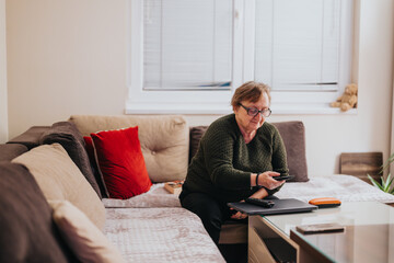 An elderly woman seated on a soft couch adjusting a remote control, depicting relaxation and...