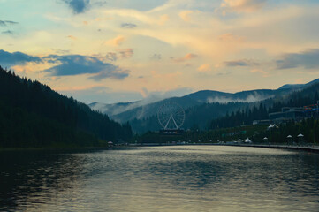 A picturesque view of the lake and mountains during sunset, Bukovel ski resort in the Carpathians. Fog drifts between the mountain peaks after rain. Ferris wheel in the background.