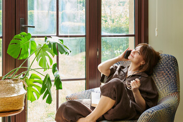 A woman relaxes in an armchair by a large window, engrossed in reading a book, surrounded by lush...