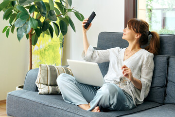 A woman comfortably works from home on a couch, using her laptop and talking on the phone,...