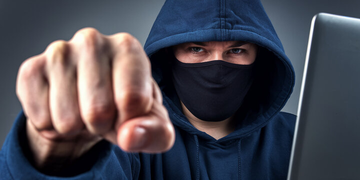 Close-up of fist and masked person in hoodie, laptop in background, symbolizing online threat, aggression, or cybercrime