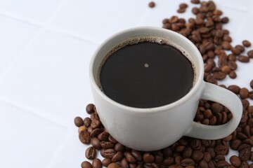 Aromatic coffee in cup and beans on white tiled table, closeup. Space for text